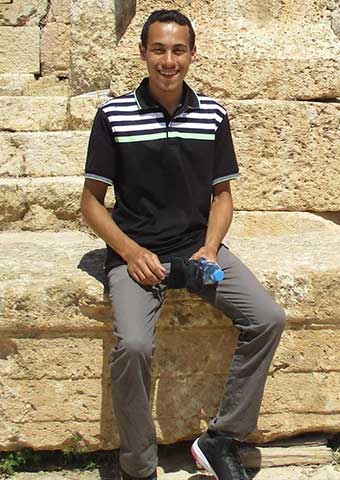 A man sitting on rocks, he has short hair and is wearing a striped polo and gray jeans