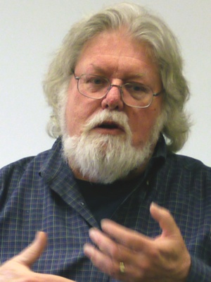 man with shoulder-length hair and beard talking, he is wearing a dark blue button-up