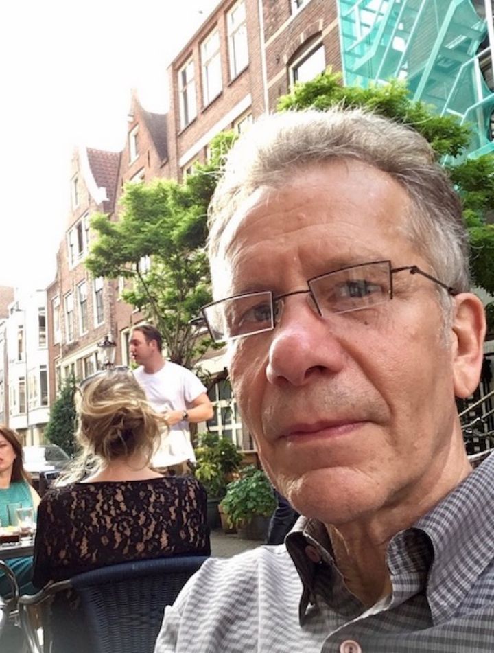 Man with glasses wearing a grey checkered shirt sitting down at a table in front of trees and brown brick buildings 