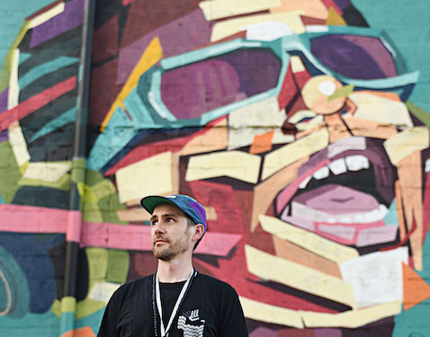 side of a building with a colorful mural with a man wearing a black tshirt and hat standing in front of it