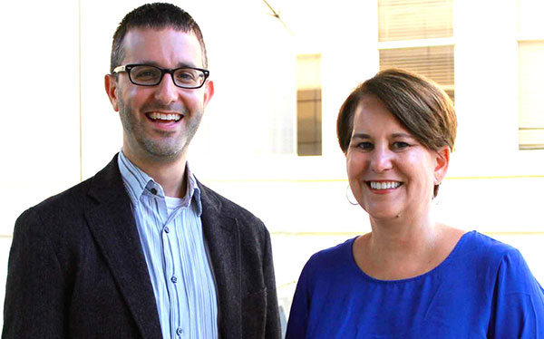 a man wearing a black suit jacket with a blue shirt and glasses and a woman with short hair wearing a blue shirt  