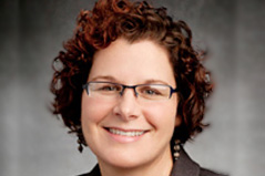 a women with curly hair wearing glasses and a gray blazer with a black shirt and a necklace