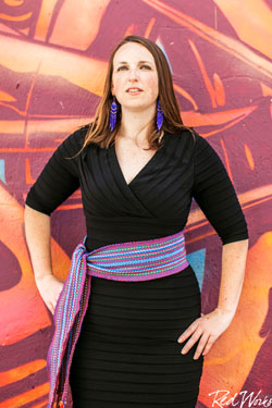 picture of a woman with medium-length brown hair. she is wearing purple feather earrings and a black dress with a cloth tied at her waist