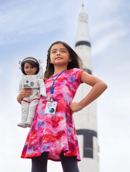 A young girl wearing a bright pink dress standing in front of a rocket while holding a doll dressed like an astronaut.