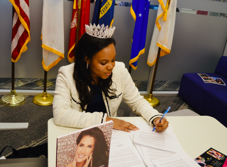 woman who's wearing a white jacket and a crown who's siting at a table and signing papers