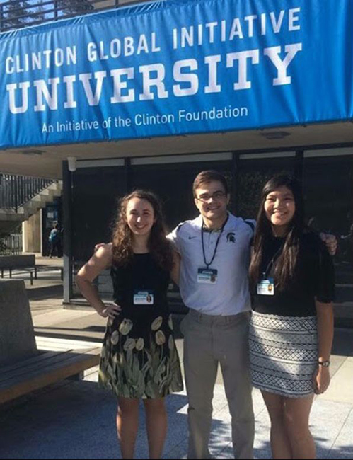 2 women and man pose for picture in front of "clinton global initiative university" sign