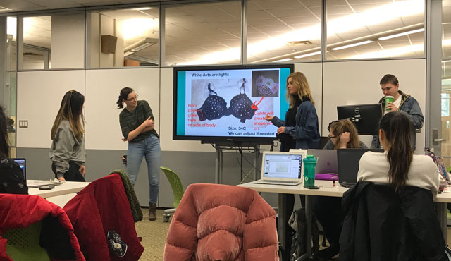 a classroom of people looking at a presentation monitor while four students present