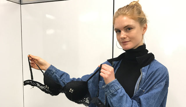 woman with a blonde bun and wearing a black turtleneck and denim jacket, she is holding up a black bra prototype