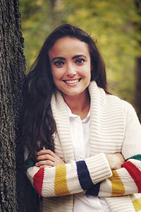 woman with long brown hair who's wearing a cream sweater and leaning against a tree.