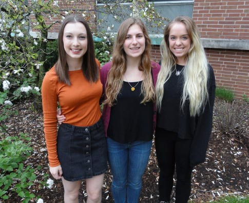 three girls one wearing an orange shirt and jean skirt, the middle girl wearing a black shirt and pink cardigan, and the third girl is wearing a black long sleeve shirt 