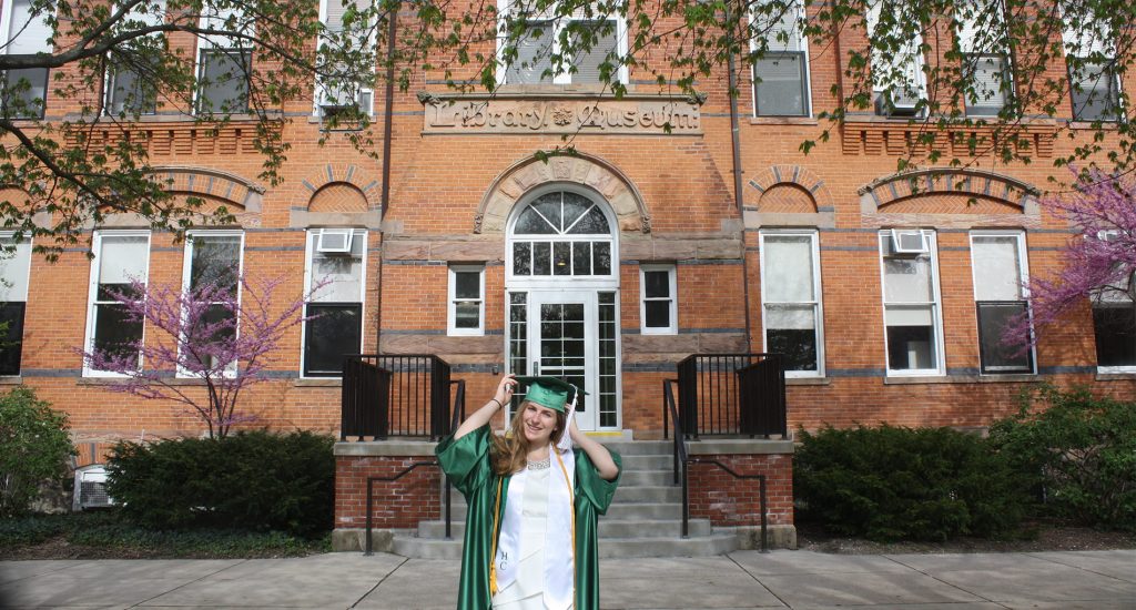 a girl standing in a green cap and gown standing in front of a brick building 