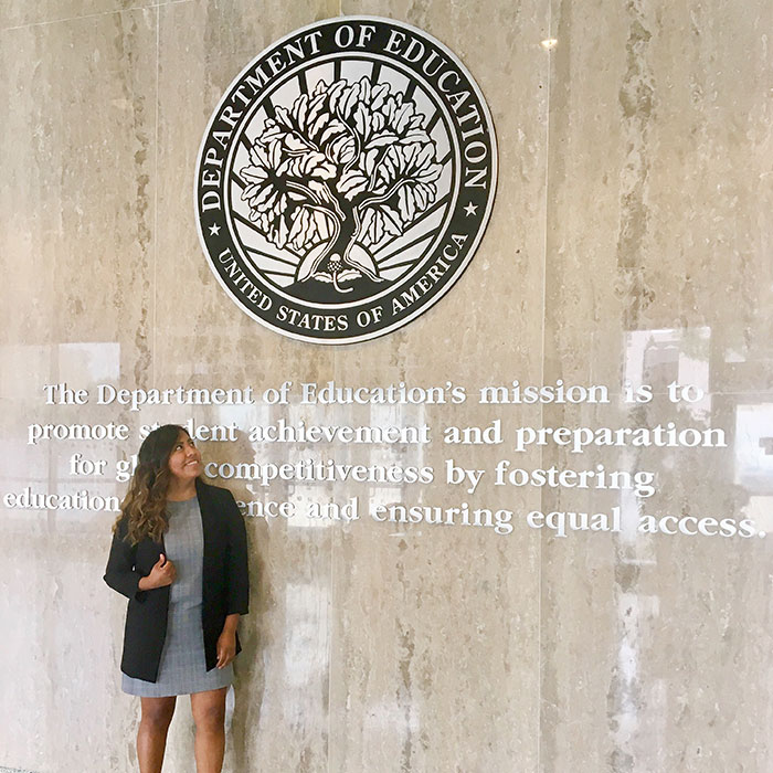 woman with brown hair and wearing a black checkered dress and black blazer posing next to the MSU department of education emblem