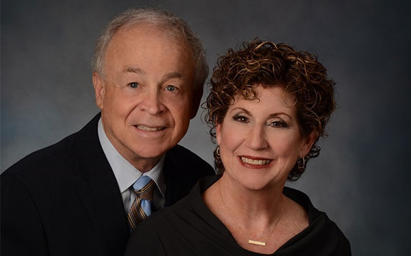 portrait of a woman and man, the man(left) has light hair and is wearing a suit, tie, and blazer. the woman(right) has short, curly, dark hair and is wearing a black top
