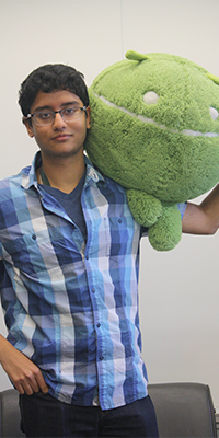 portrait of a man holding an android plush in his left arm, he has dark hair, glasses, and is wearing a plaid button-up