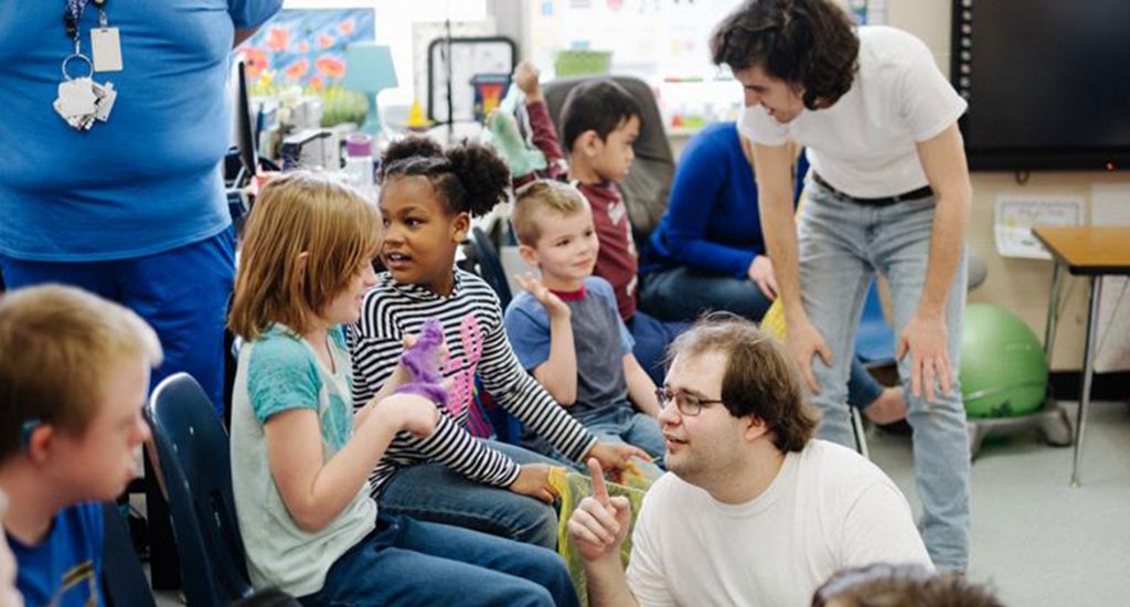 A man in a white shirt is kneeling and talking to a child that's sitting in a chair. Behind him, a man also wearing a white shirt is bending over to talk to a child sitting down.