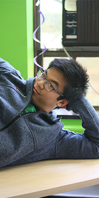 portrait of a man posing on a desk while laying on his side. he has black hair and rectangular glasses, he is wearing a green shirt and a zip-up sweater