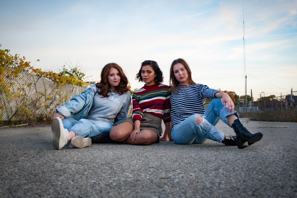 three girls sitting together in a road in front of a vine covered fence and blue sky