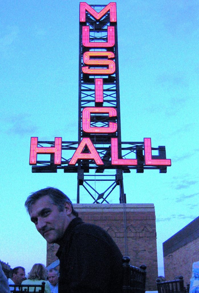 man wearing a black shirt sitting in front of large neon sign that says music hall