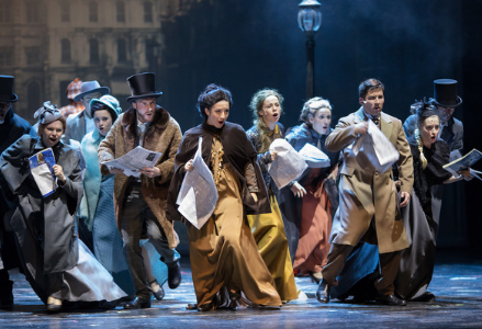 a group of six women and four men on a stage, who are all wearing neutral, old fashioned clothing who are running forward and look as if they are singing