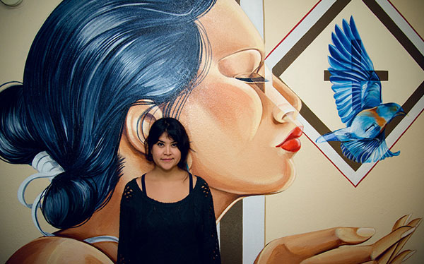 woman standing in front of painting of woman with blue hair and blue bird flying out of her hand