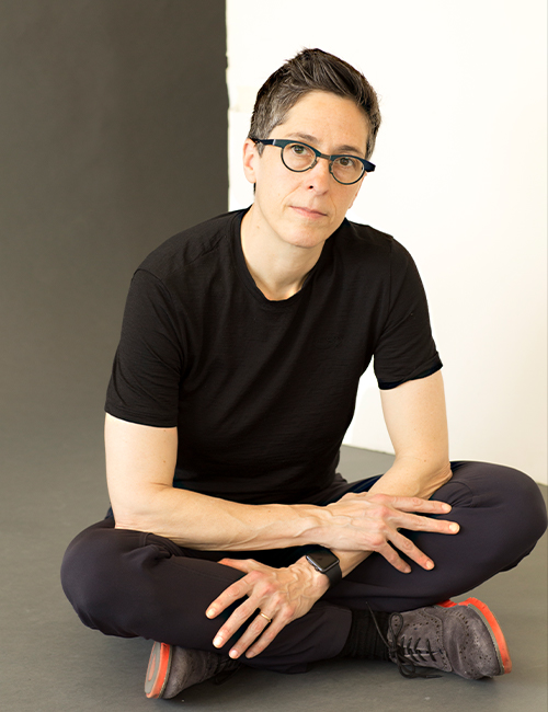 Alison Bechdel wearing black pants and a black shirt sitting on the floor