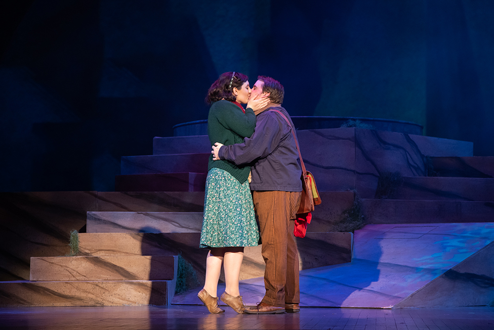 a woman wearing a green dress and a man wearing a brown outfit and hat kissing on stage