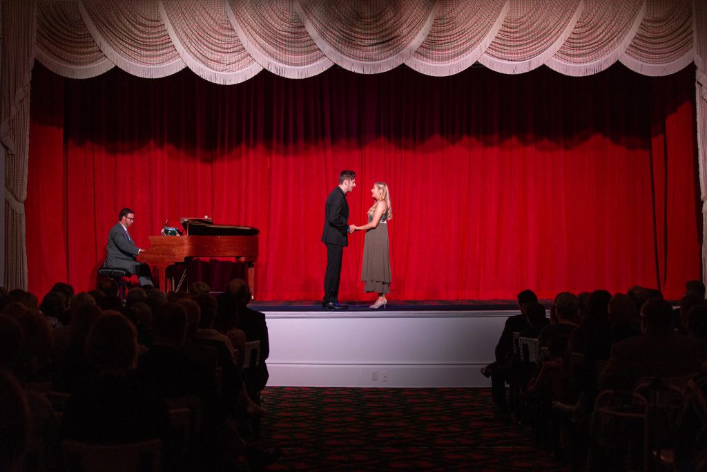 man and woman holding hands on a stage with a red backdrop in front of an audience 