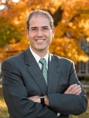 man wearing glasses and a gray suit and green tie with his arms crossed smiling at the camera
