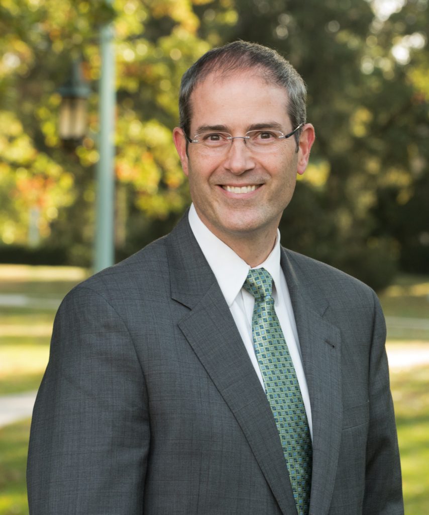 Dean Christopher Long in a grey suit and a green tie smiling at the camera