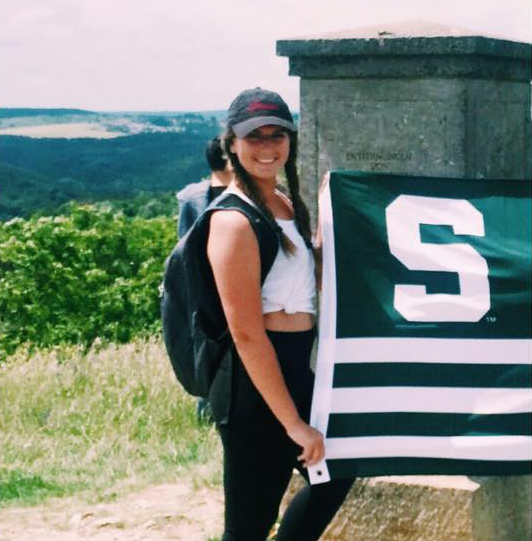 a girl with her hair braided wearing a hat a white tank black leggings and a backpack holding an MSU flag