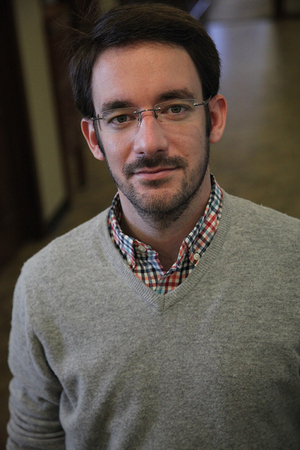portrait of a man with short dark brown hair, he is wearing glasses and a gray sweater over a plaid button-up