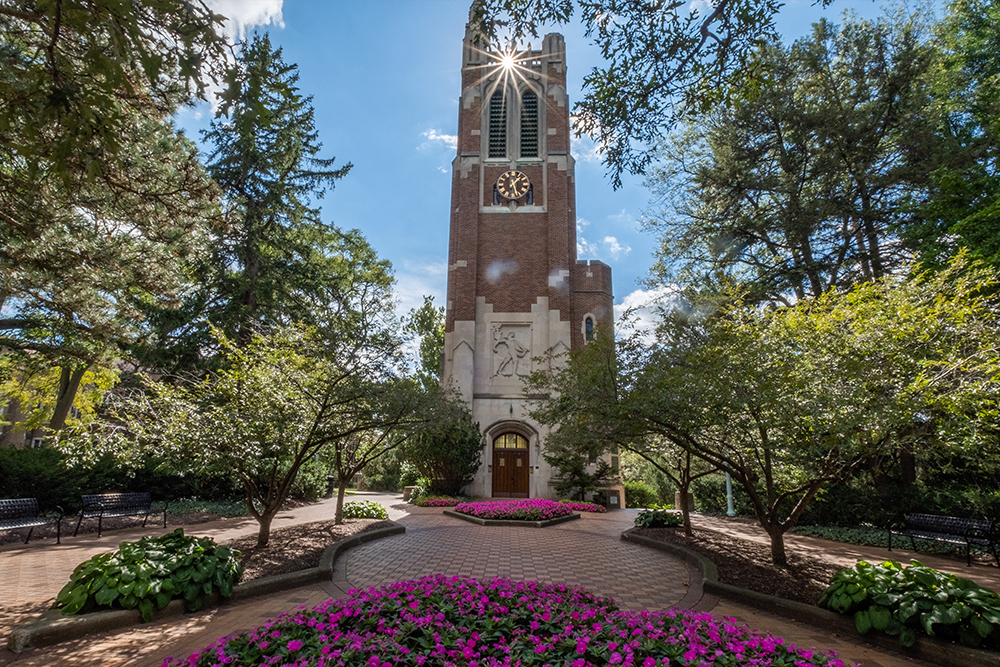 red brick bell tower in the center of a plaza with flowers blooming and trees surrounding