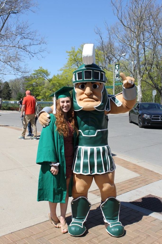 Woman in a green cap and gown standing outside next to Sparty 