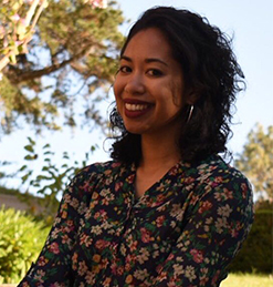 portrait of a smiling woman with shoulder-length, curly, black hair. she is wearing a long-sleeved, floral patterned button-up and hoop earrings