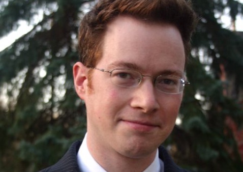 a headshot of a man with short hair wearing glasses, and black suit, and a pink tie