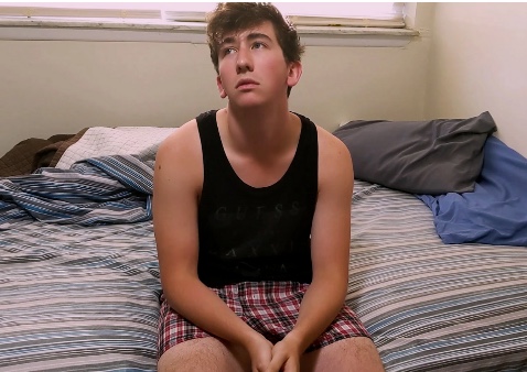 a boy sitting on a bed in a black tank and boxer shorts (a character in a film)