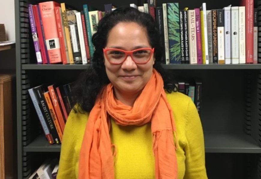 woman wearing red glasses and a bright yellow shirt with a coral scarf in front of a bookshelf