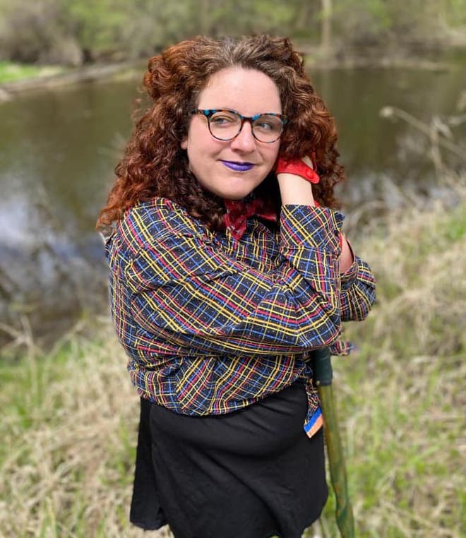 a woman with long curly hair wearing glasses and a plaid shirt with a black skirt 