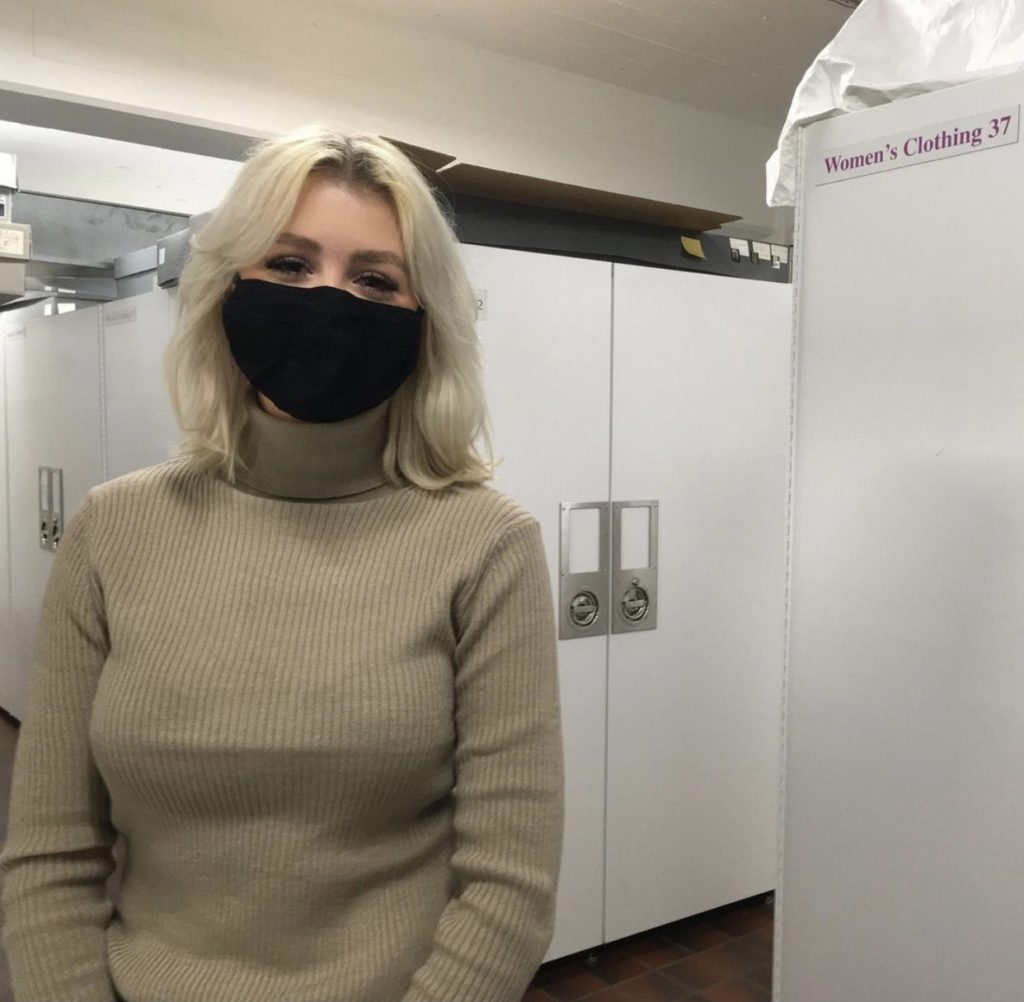 woman wearing a tan turtleneck and black mask sting in a room with white cabinets