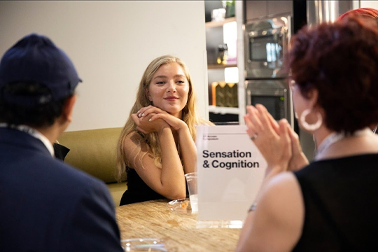 Woman with blonde hair sitting at a table talking to two other people