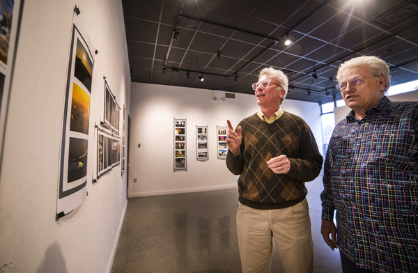 Photo of two men standing side by side in an art gallery. The man on the left is pointing at a photo on the wall.