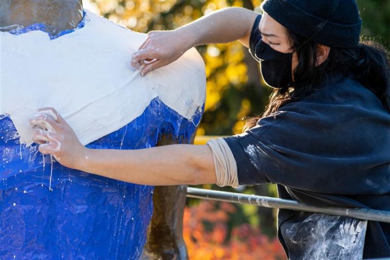 Photo of a person taking a silicone cast of a bronze Spartan statue. They are wearing a navy blue hat, black face mask, and a navy blue sweatshirt.