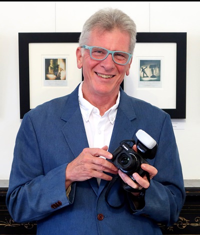A smiling man with gray hair wearing light blue glasses and holding a camera. Behind him are two framed photos. 