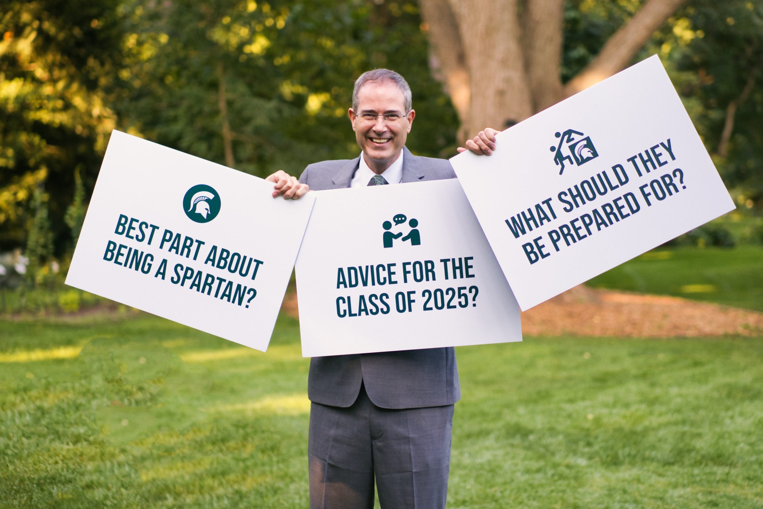 Man wearing a gray suit and holding up three white poster board signs. The text is "Best part about being a Spartan?" and "Advice for the class of 2025?" and "What should they be prepared for?"