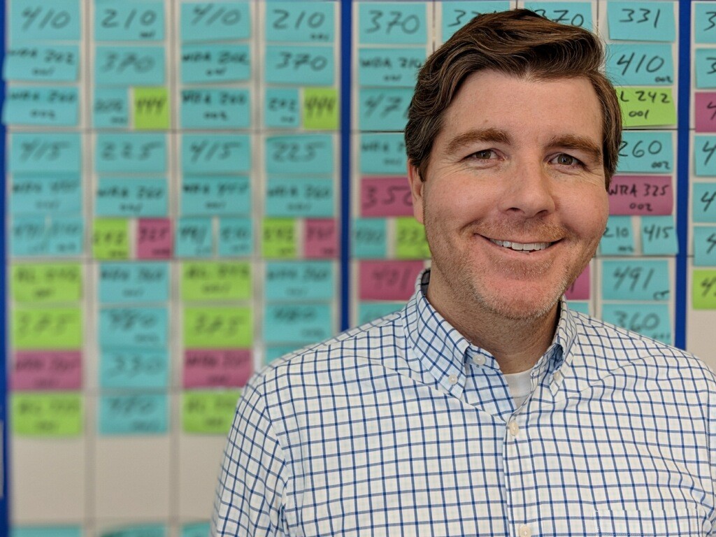 Man with reddish-brown hair is smiling. Behind him are blue, green, and pink sticky notes with numbers on them.