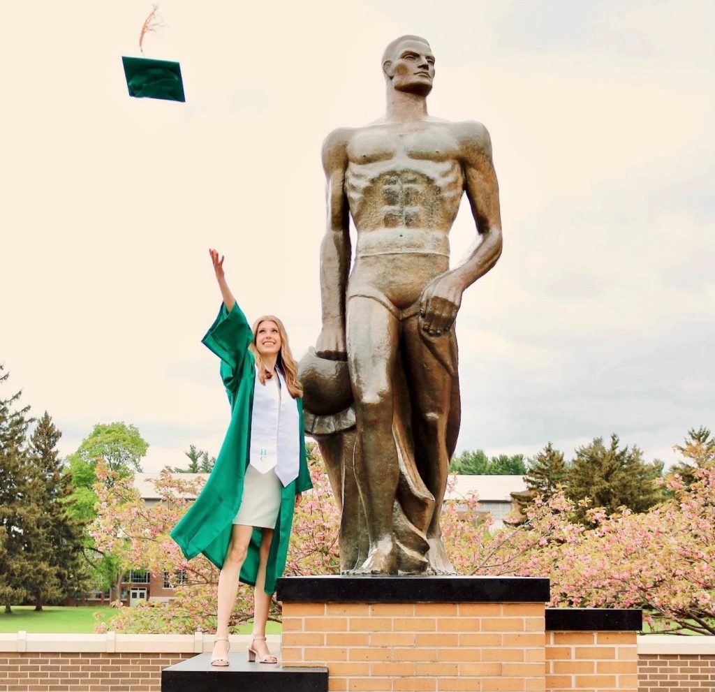 A woman with light hair wearing a dress and green graduation gown is tossing her green graduation cap. She is standing on the platform to the left of statue of a man.