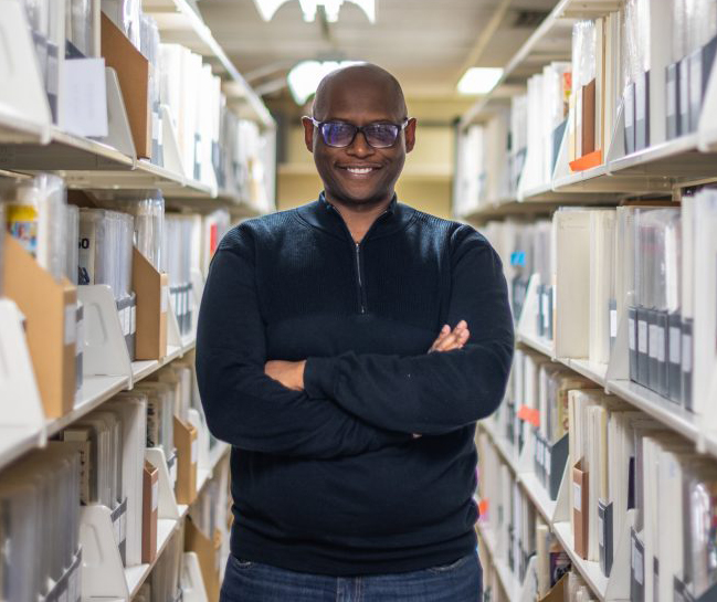 Man with glasses is standing with his arms crossed over his chest. He is standing between two bookshelves in a library.