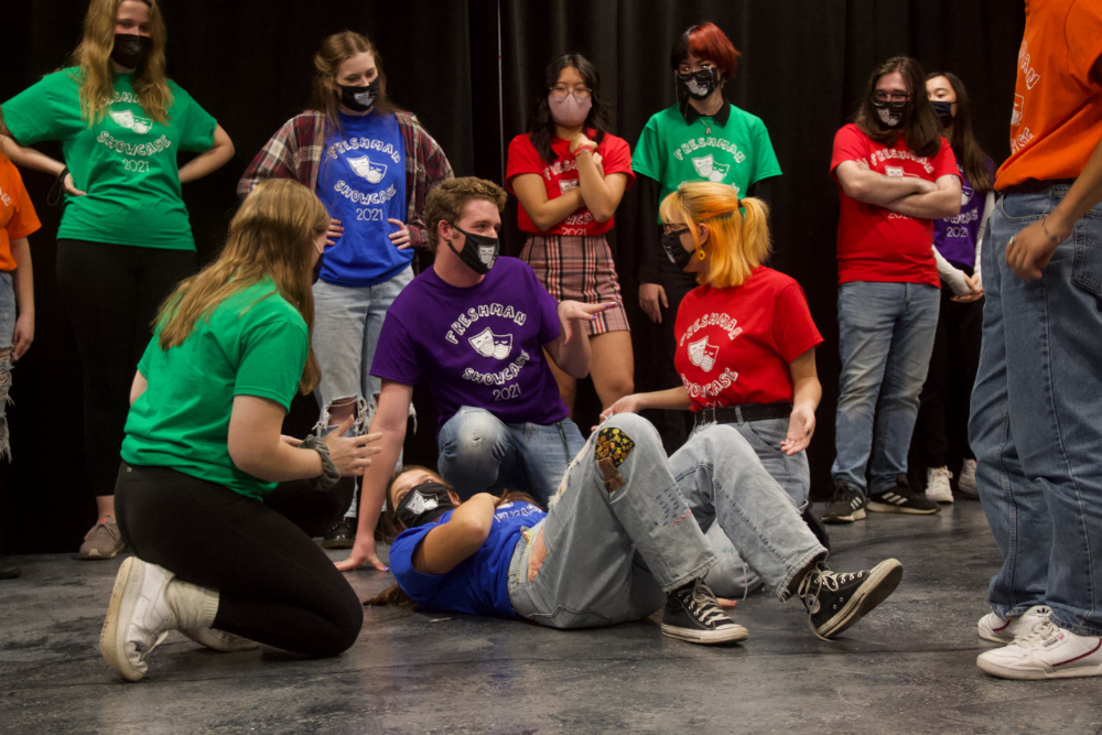 Students rehearse for the improv comedy show. One student in a blue shirt lies on the ground, surrounded by three kneeling students. Other students watch in the background