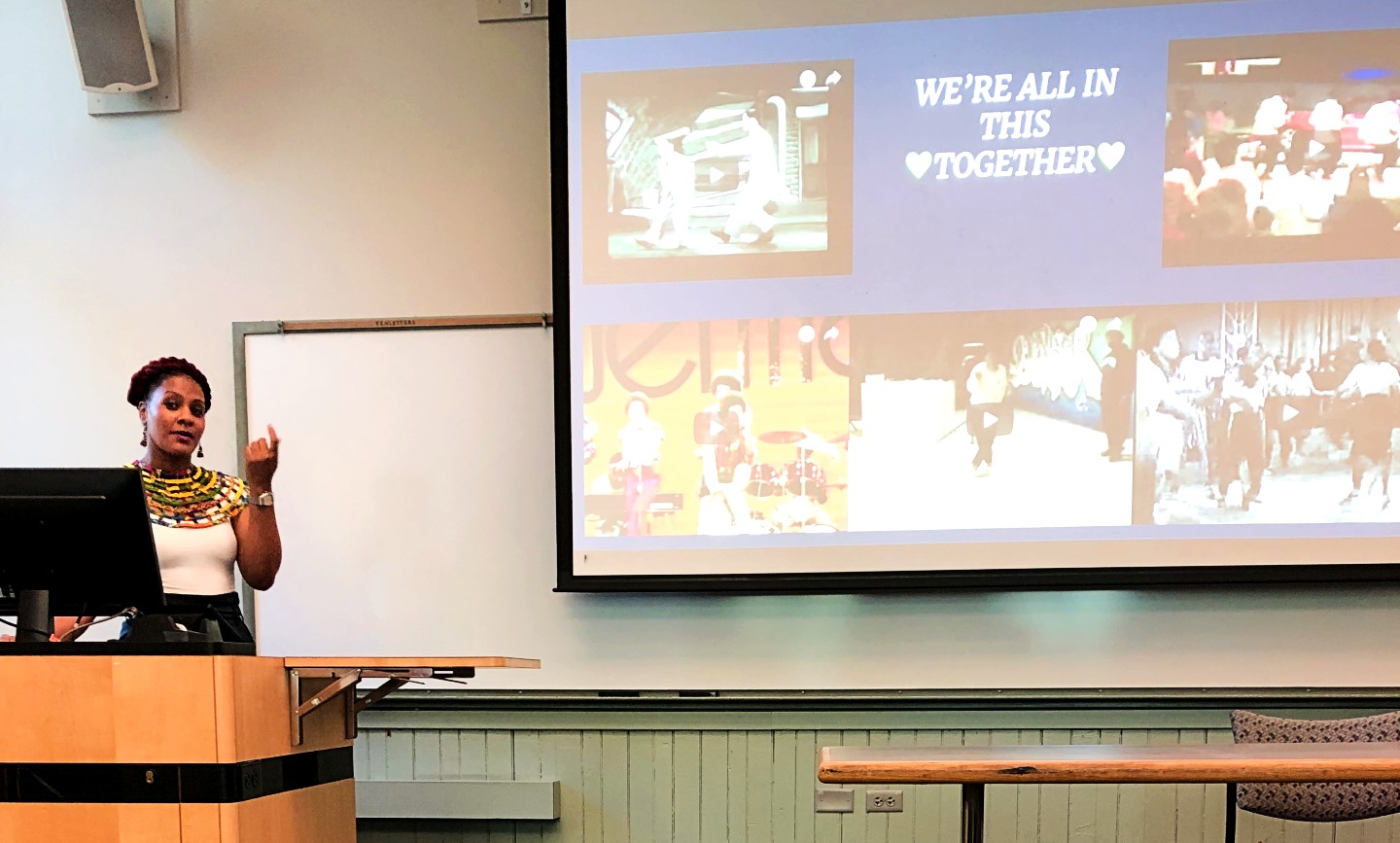 A woman at a computer stand in the middle of talking next to a projector screen displaying photos and the text, "We're all in this together" with a heart on each side of the word "together".