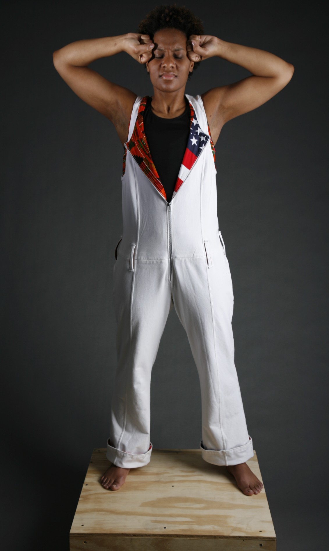 A professional photo of a woman standing tall with eyes closed and arms raised with elbows bent and fists pressed against her temples. She is wearing a white jumpsuit with the front unzipping to show the American flag on the inside.
